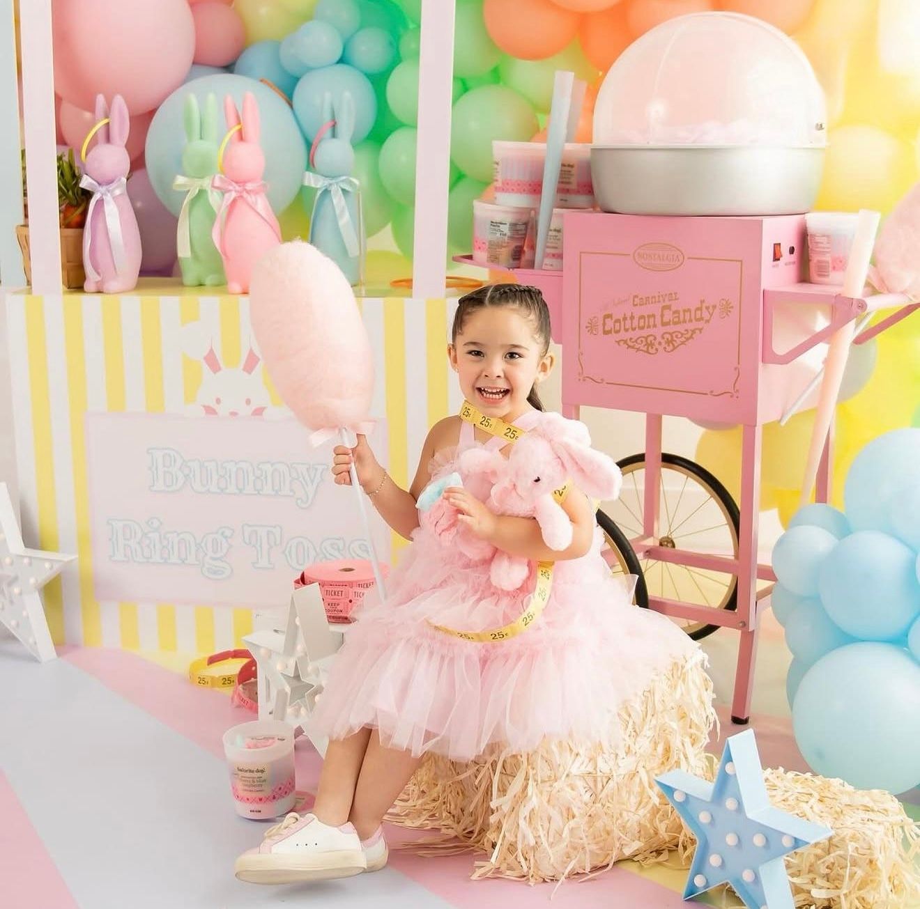 A smiling girl in a pink tutu holds cotton candy beside a vintage pink cotton candy machine at a pastel bunny-themed party.