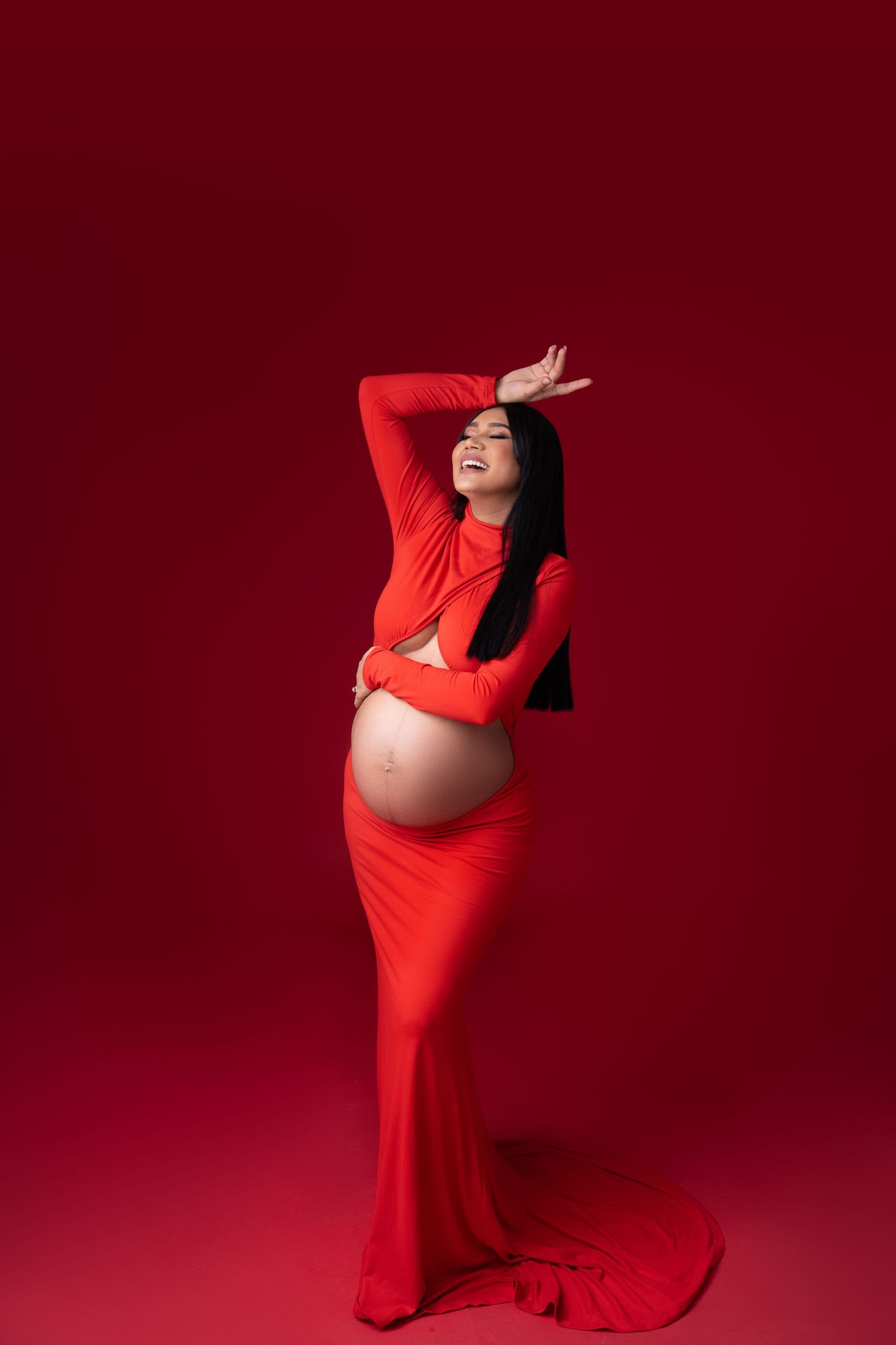 Elegant maternity photo featuring a flowing red dress against a deep burgundy background in a dramatic studio pose.