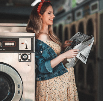 Woman in floral dress and denim jacket smiles while doing laundry at a laundromat with vintage washing machines.