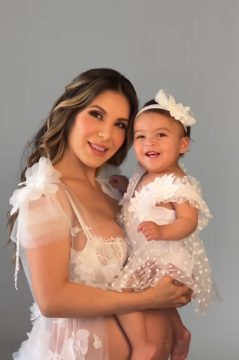 Mother and baby in matching white lace dresses pose together for a portrait against a gray background.