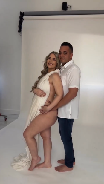 Couple embracing during intimate maternity photoshoot in white studio setting.