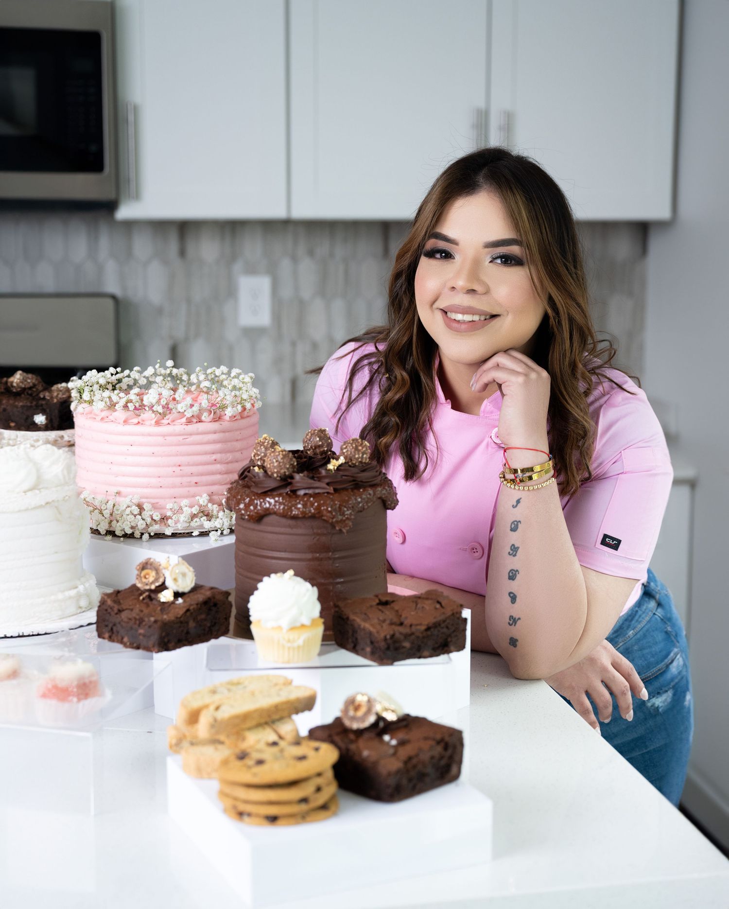Baker in pink sweater displays homemade desserts including chocolate cake, pink cake, brownies and cookies in modern kitchen.
