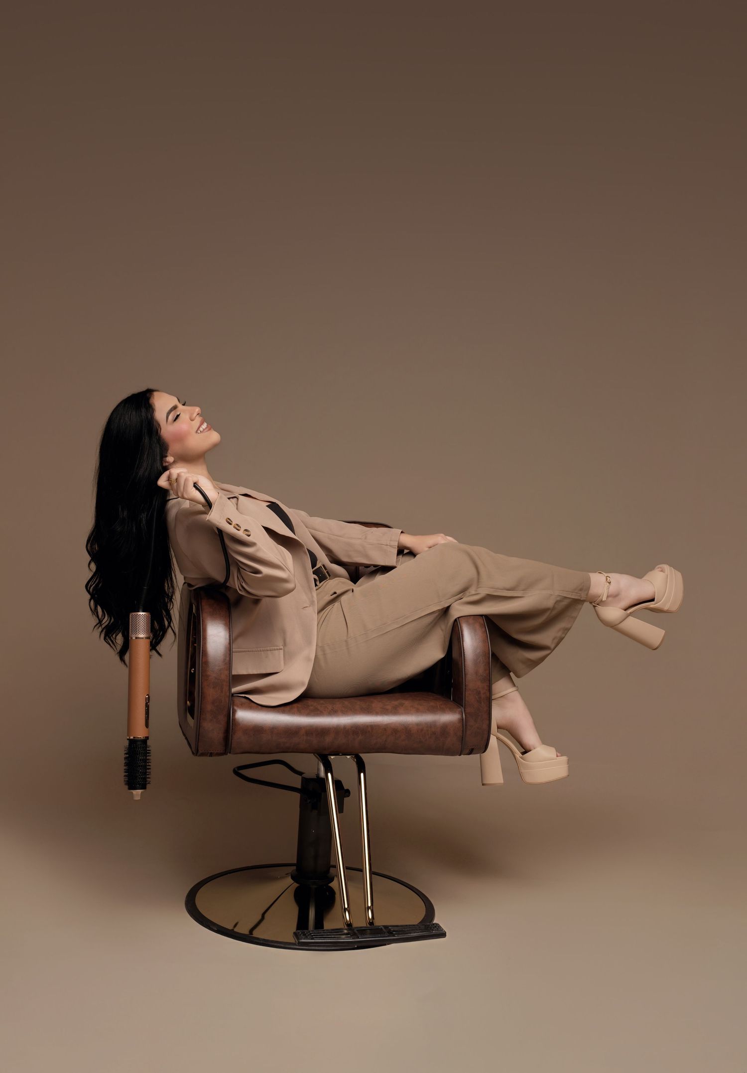 Person in beige outfit lying horizontally across brown leather chairs in artistic fashion photoshoot series.