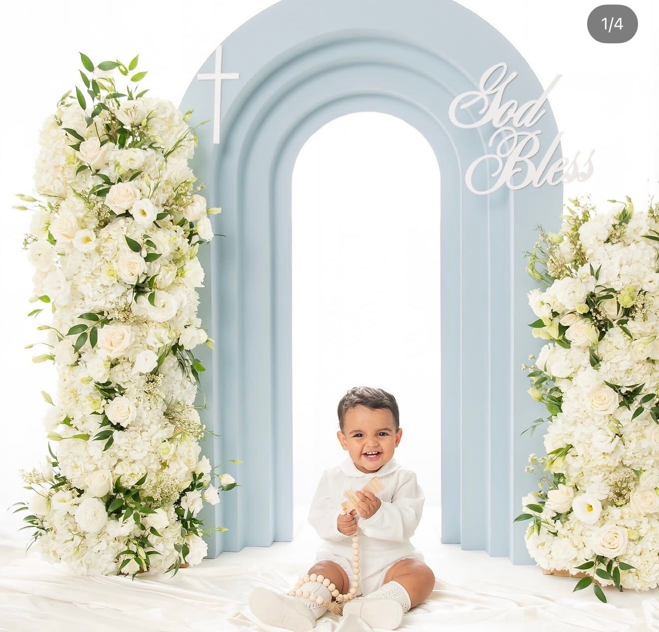 A smiling baby in white outfit sits before an elegant arch backdrop with white floral arrangements and God Bless sign.