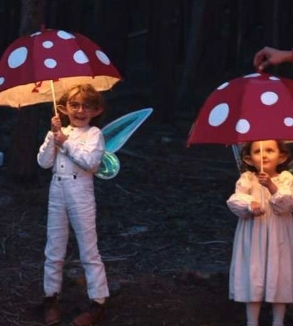 Two children in white costumes holding red and white polka dot mushroom umbrellas in a forest setting.