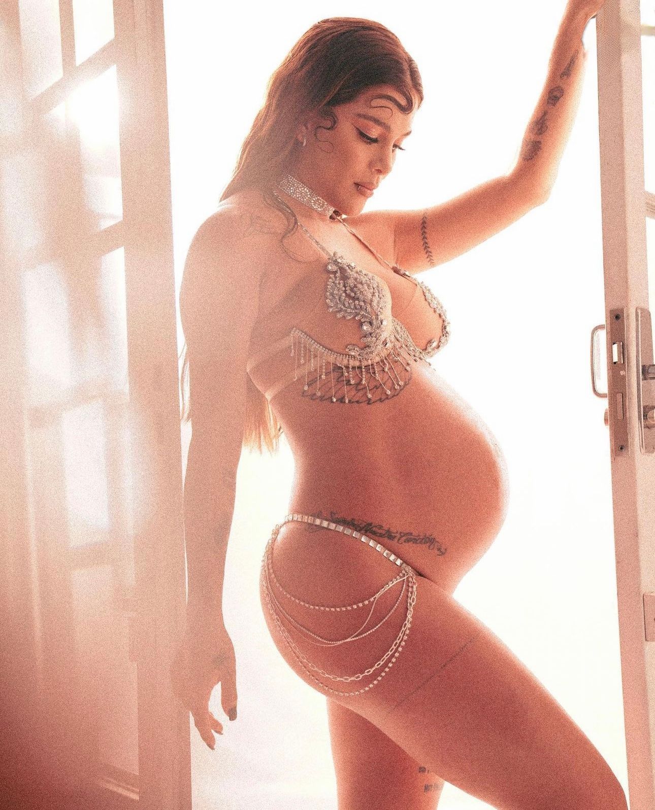 Pregnant woman posing in sparkly crystal bra and chain body jewelry in soft backlit doorway.