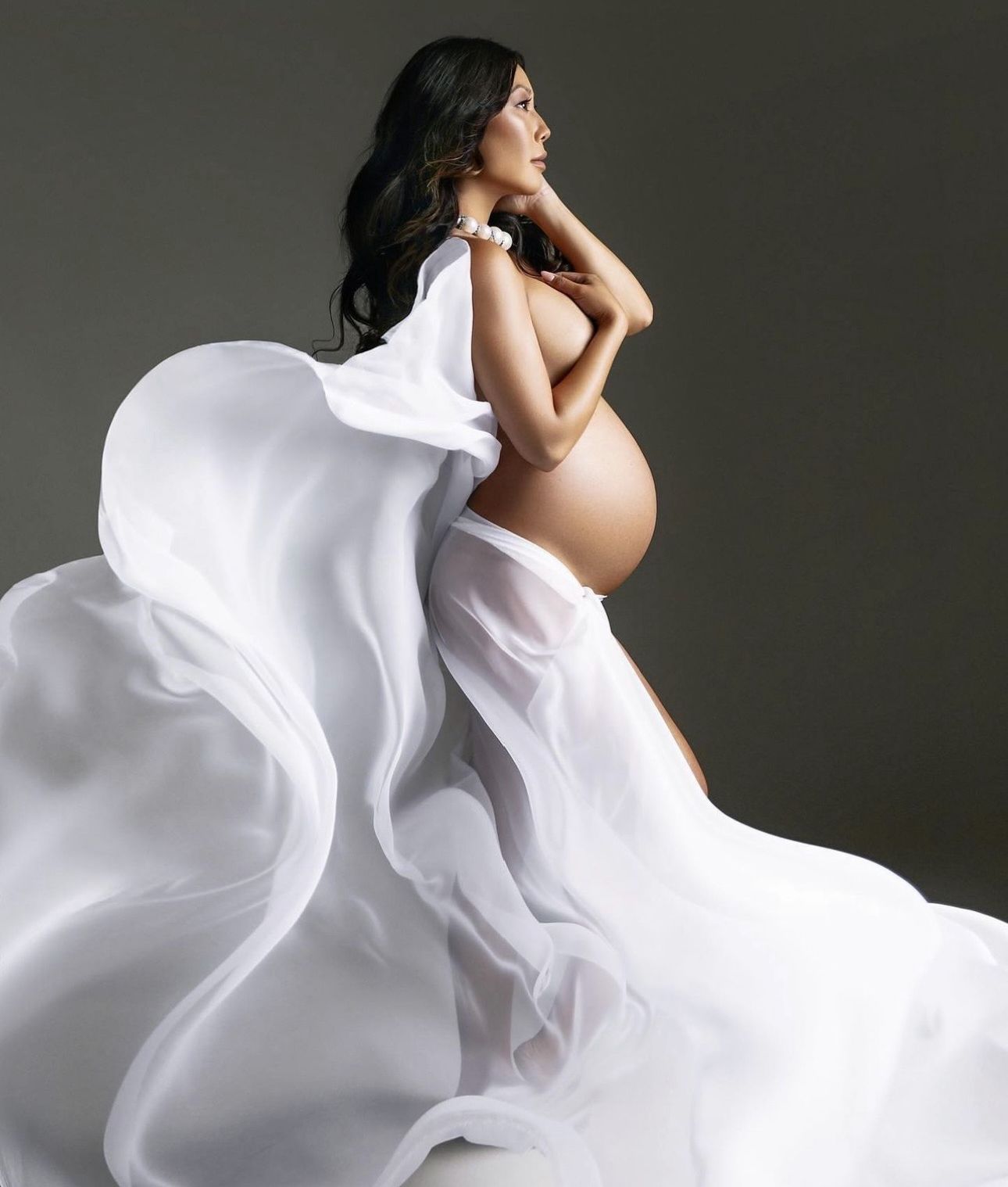 Pregnant woman draped in flowing white fabric, showcasing her baby bump in an elegant maternity photography studio portrait.