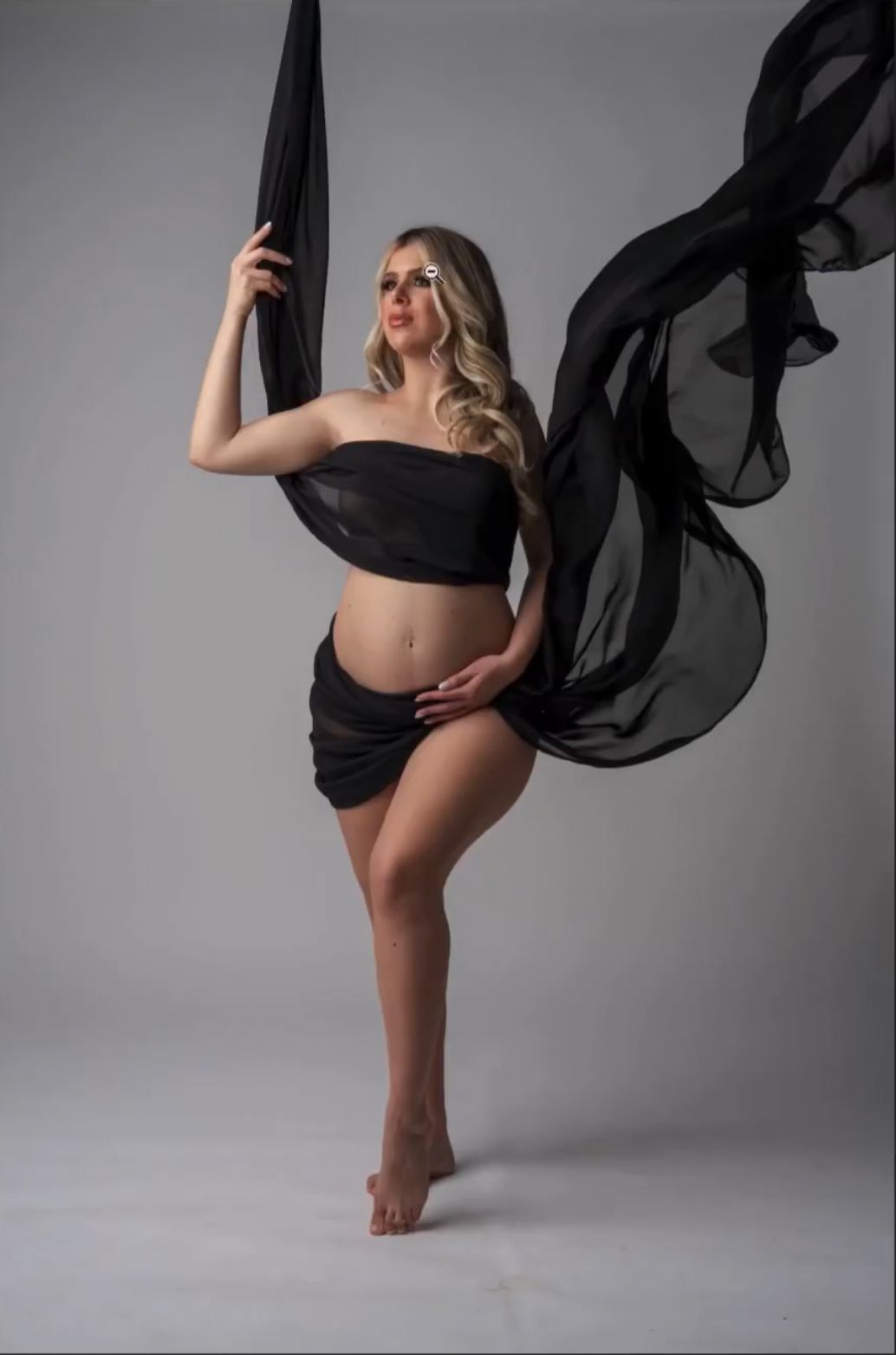 Pregnant woman in black bandeau and draped skirt poses gracefully with flowing black fabric in a studio maternity photo.