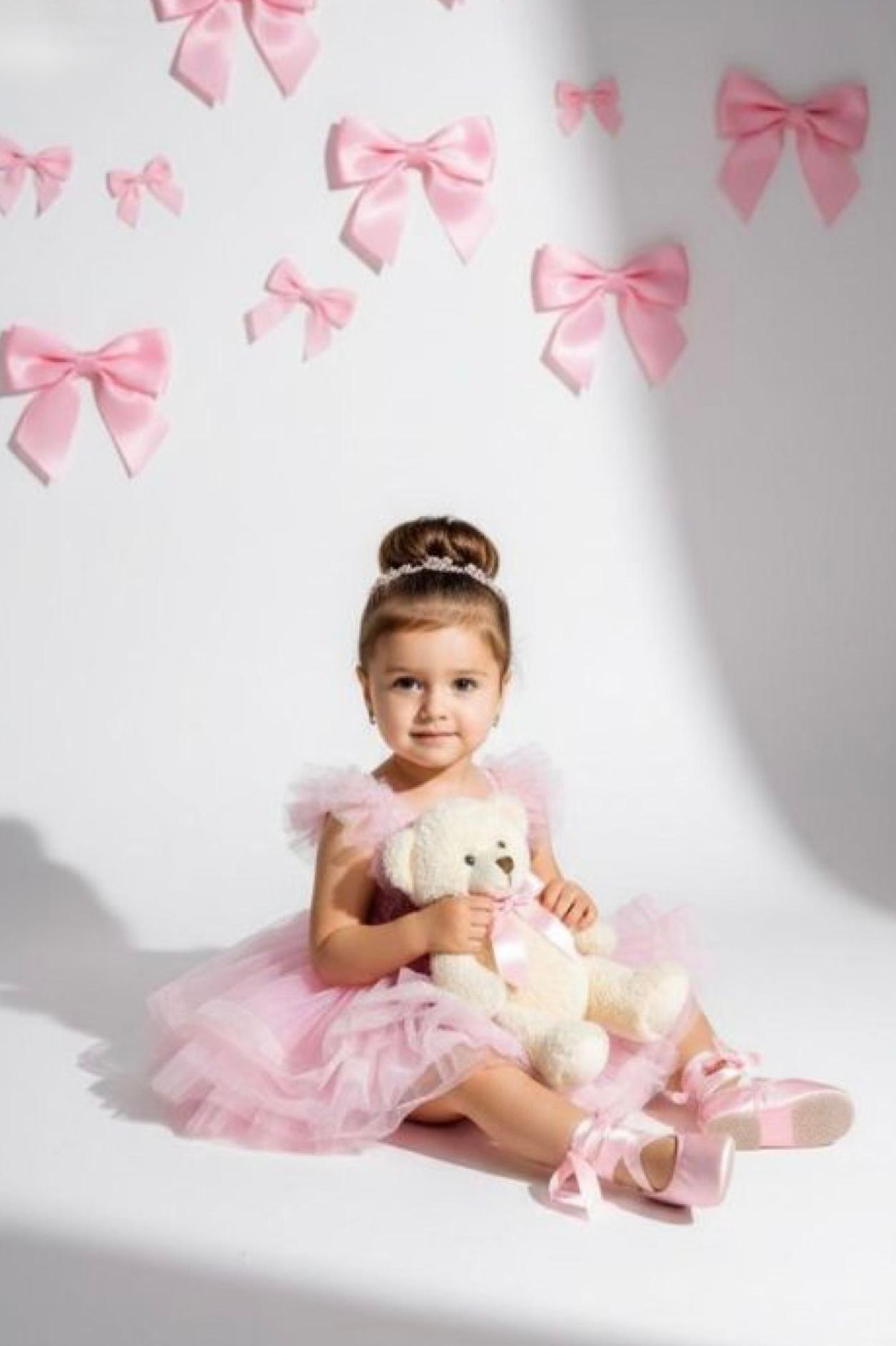Toddler girl in pink tutu dress holding teddy bear, sitting against white wall decorated with pink satin bows.