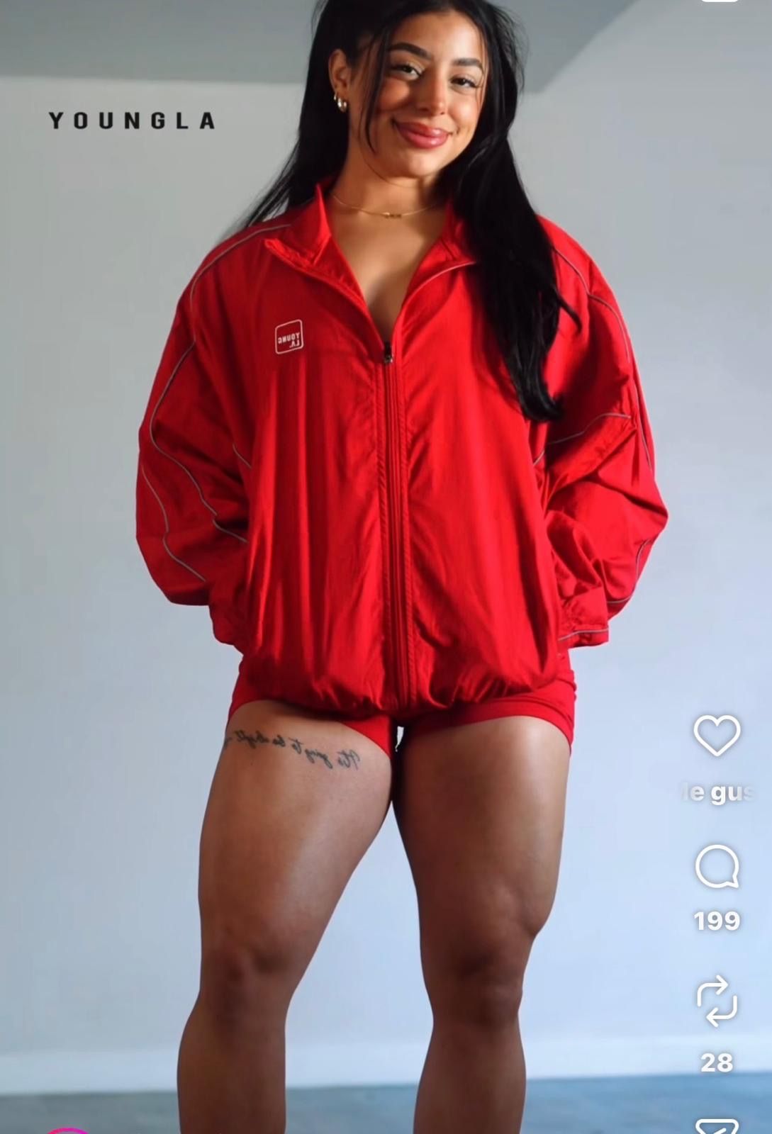 Muscular woman in red oversized YoungLA track jacket and shorts showing athletic legs with thigh tattoo.