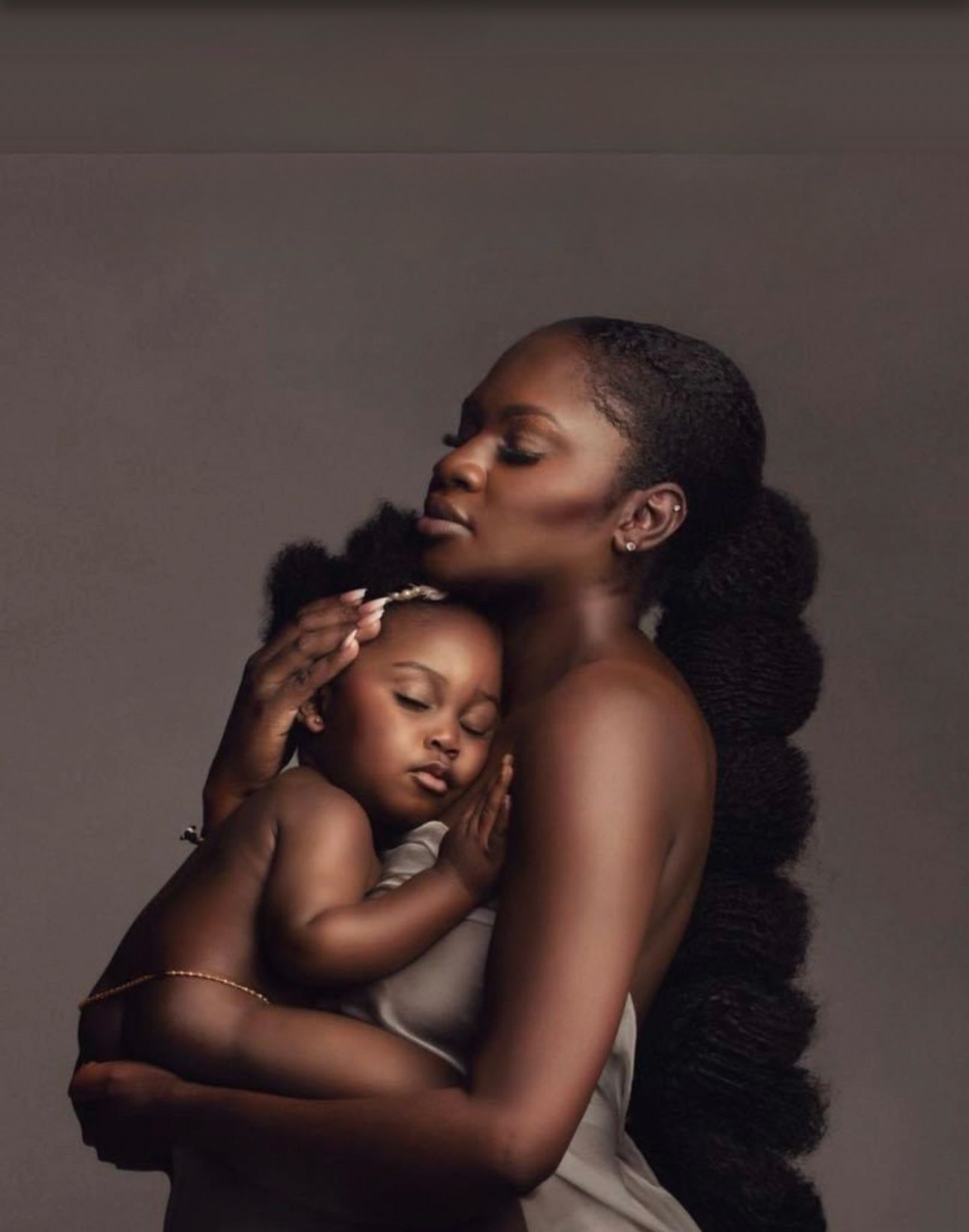 A mother tenderly holds her sleeping baby close, both dressed simply, in a warm studio portrait celebrating motherhood.