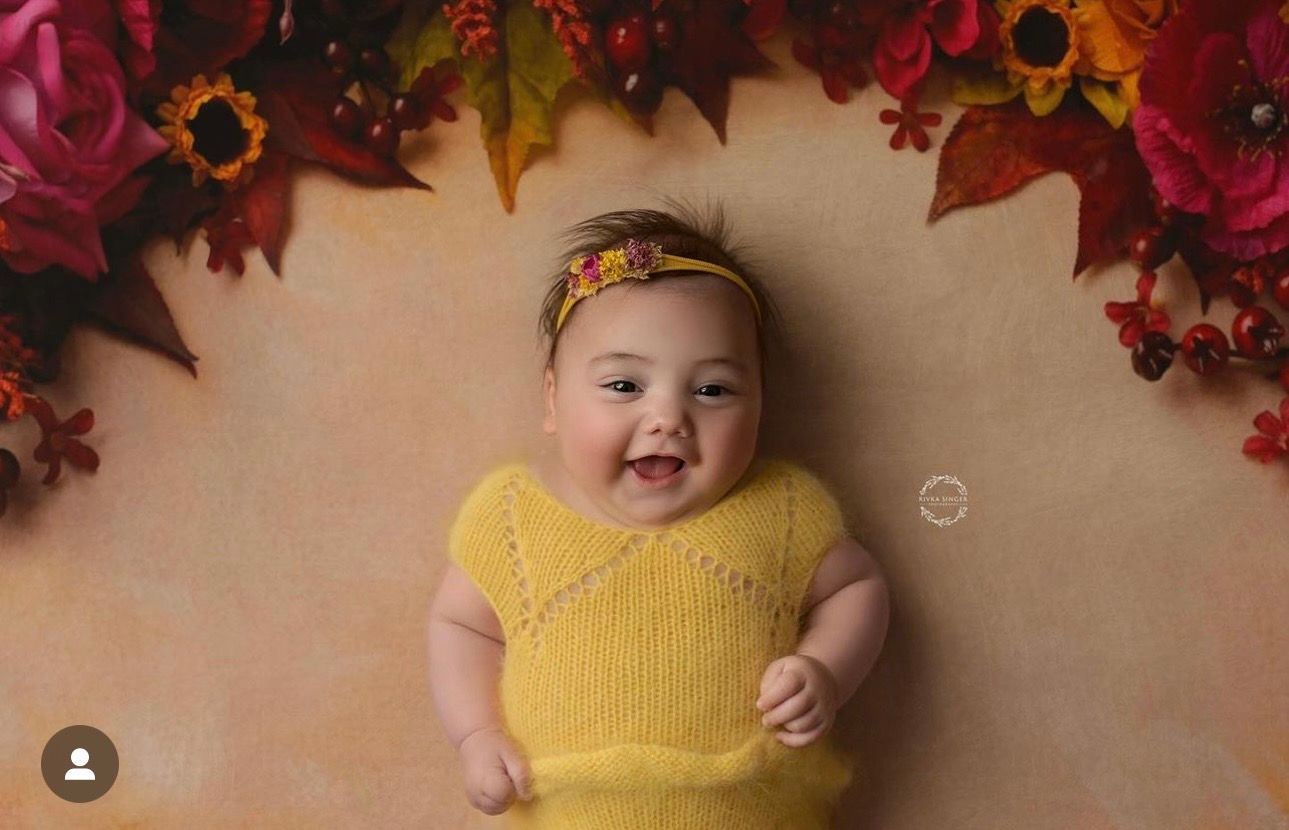 Smiling baby in yellow knit outfit with floral headband, lying on autumn backdrop with colorful fall leaves and flowers.