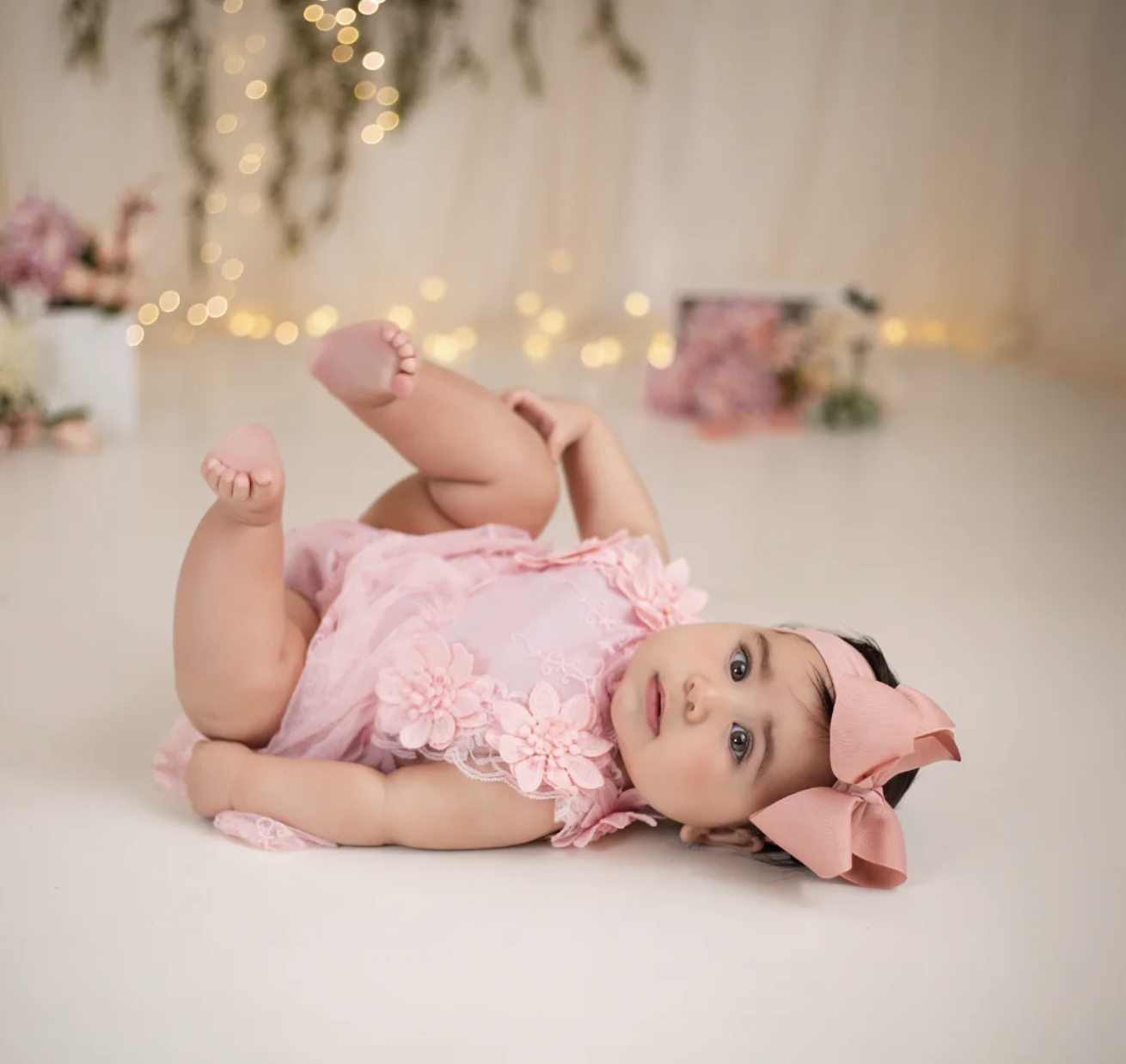 Adorable baby girl in a pink floral romper and bow headband lying on a white floor with bokeh lights behind her.