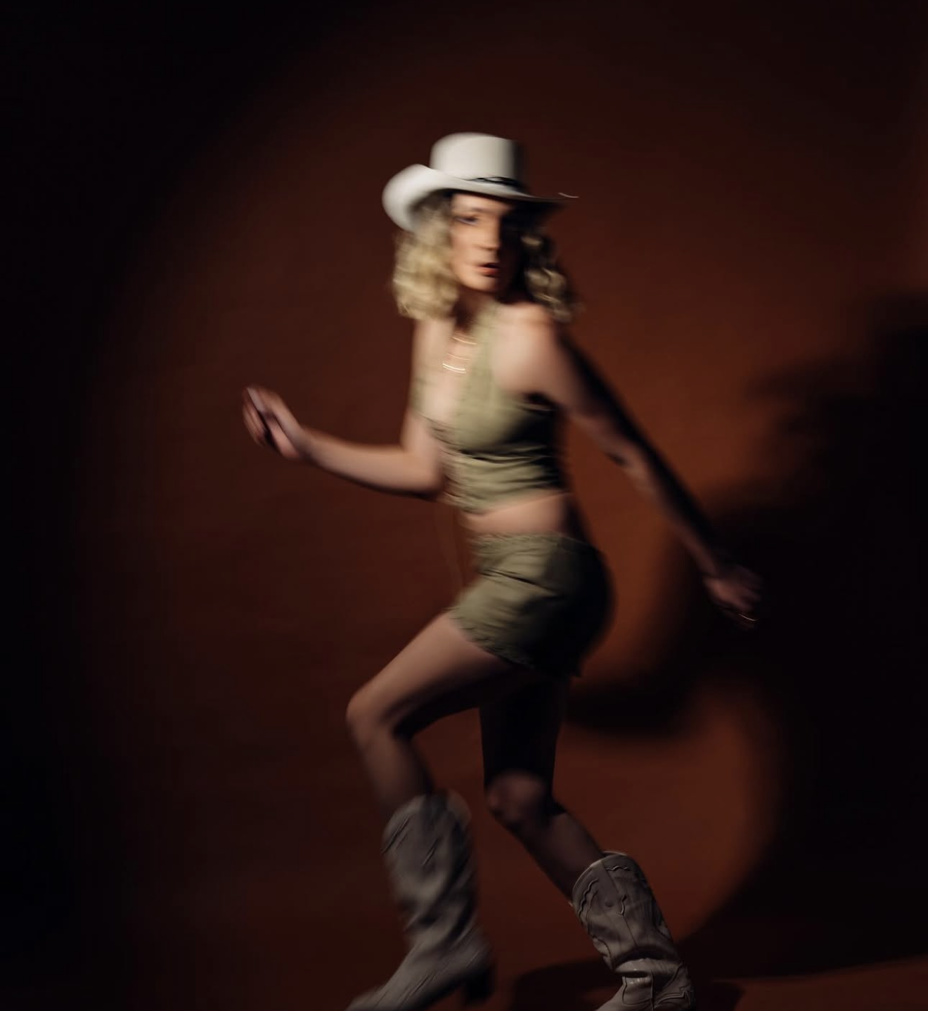 A blonde woman in cowboy hat, green crop top, shorts, and boots poses dynamically against a dark brown background.