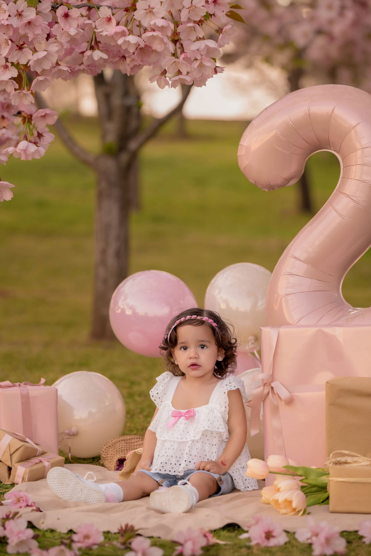 Toddler girl in white top sits among pink gifts and balloons at a cherry blossom tree 2nd birthday photo shoot.
