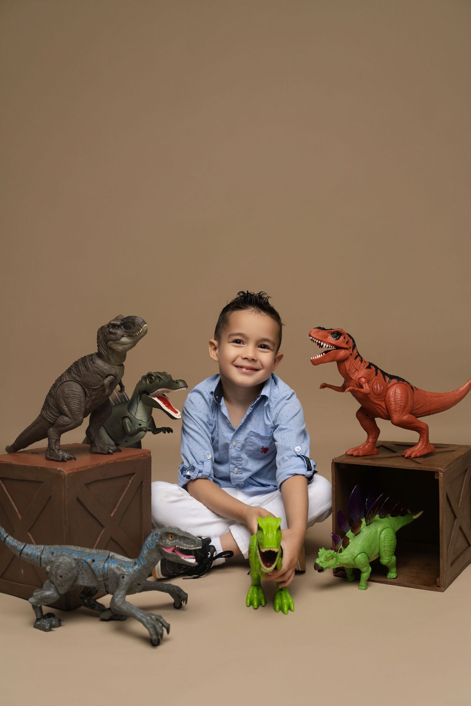 Smiling boy in blue shirt sits cross-legged surrounded by dinosaur toys on wooden boxes against beige background.