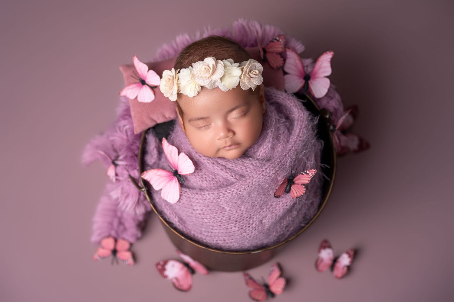 Newborn baby girl wrapped in purple swaddle inside a bucket, adorned with butterfly decorations and a floral headband.