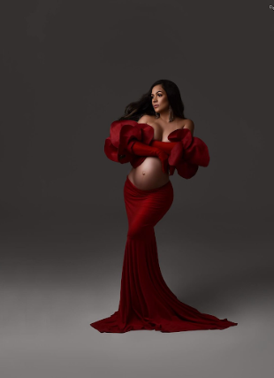 A woman in a flowing deep red velvet gown poses gracefully against a dark gray backdrop in a fashion photoshoot.