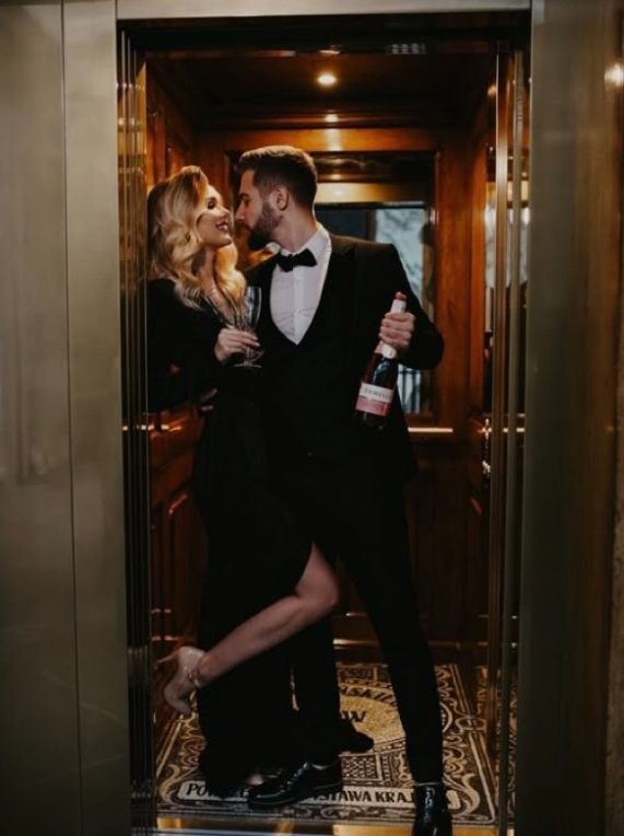 A couple in formal attire share an intimate moment in a dimly lit elevator with elegant decor.