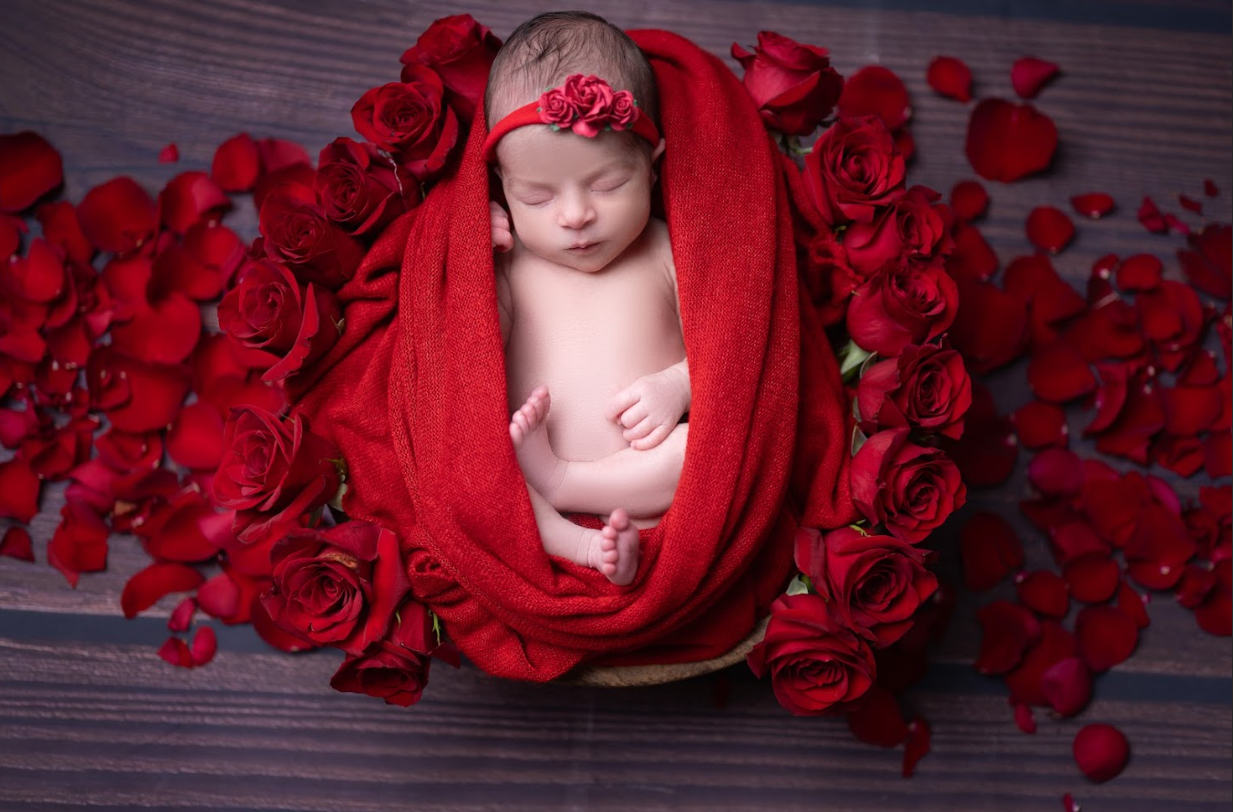 Sleeping newborn wrapped in red fabric surrounded by vibrant red roses on dark wooden background.