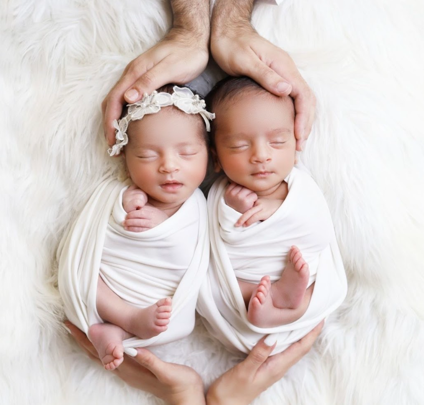 Two sleeping newborn babies swaddled in white fabric lying on a fluffy white surface with hands gently cradling them.