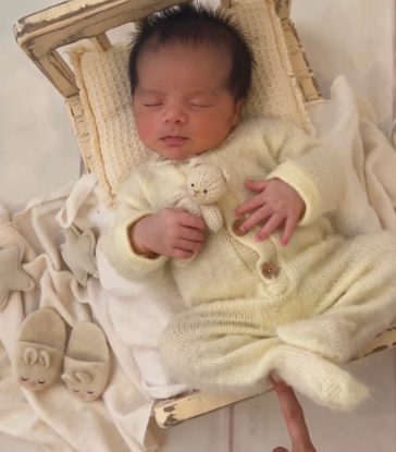 Sleeping newborn wearing a soft cream-colored sweater rests peacefully on light bedding.