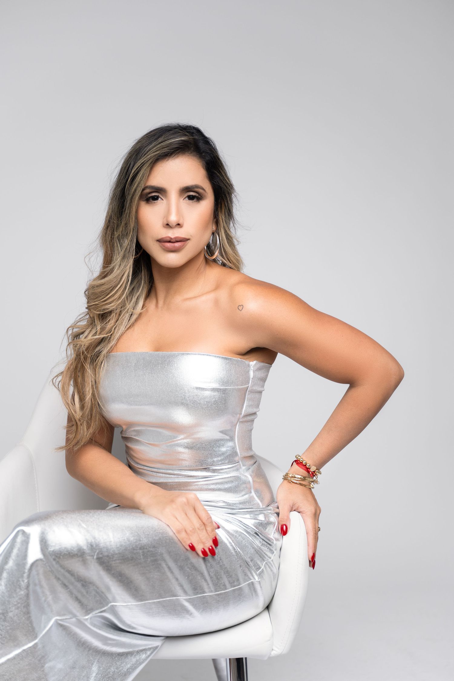 Woman with long highlighted hair poses seated on a white chair in a strapless metallic silver gown with red nails and gold bracelets.