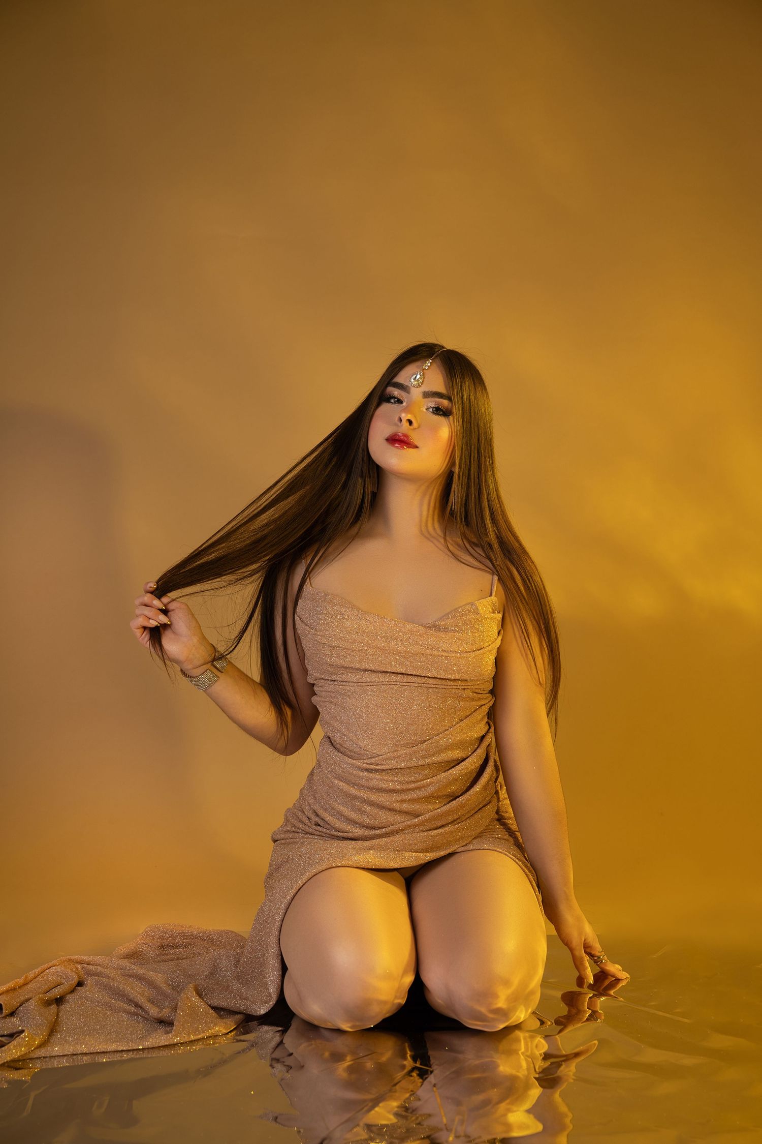 Young woman in beige dress kneeling on reflective surface with golden lighting, holding long hair, wearing forehead jewelry.