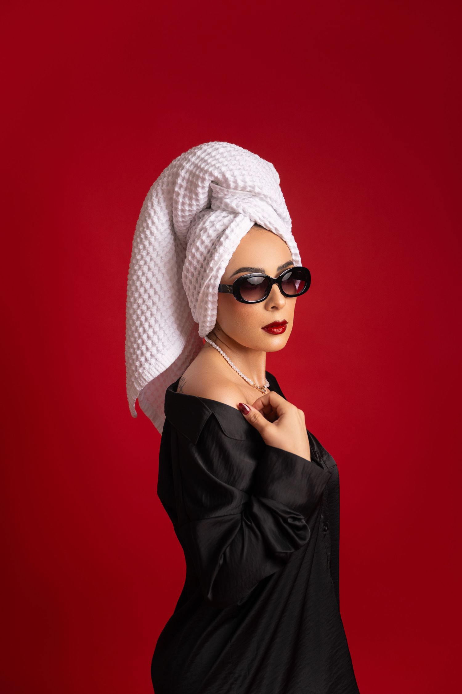 Woman in a black satin robe, sunglasses and red lipstick with a white waffle towel wrapped on her head, posing against a bold red backdrop.