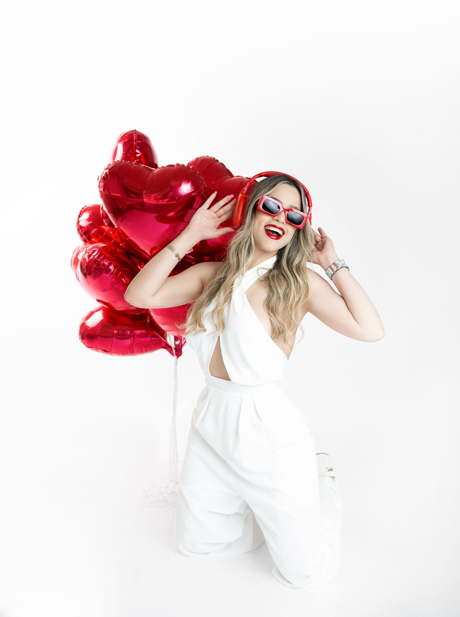 Smiling woman in a white jumpsuit, red sunglasses and red headphones kneels with a bunch of red heart-shaped balloons.