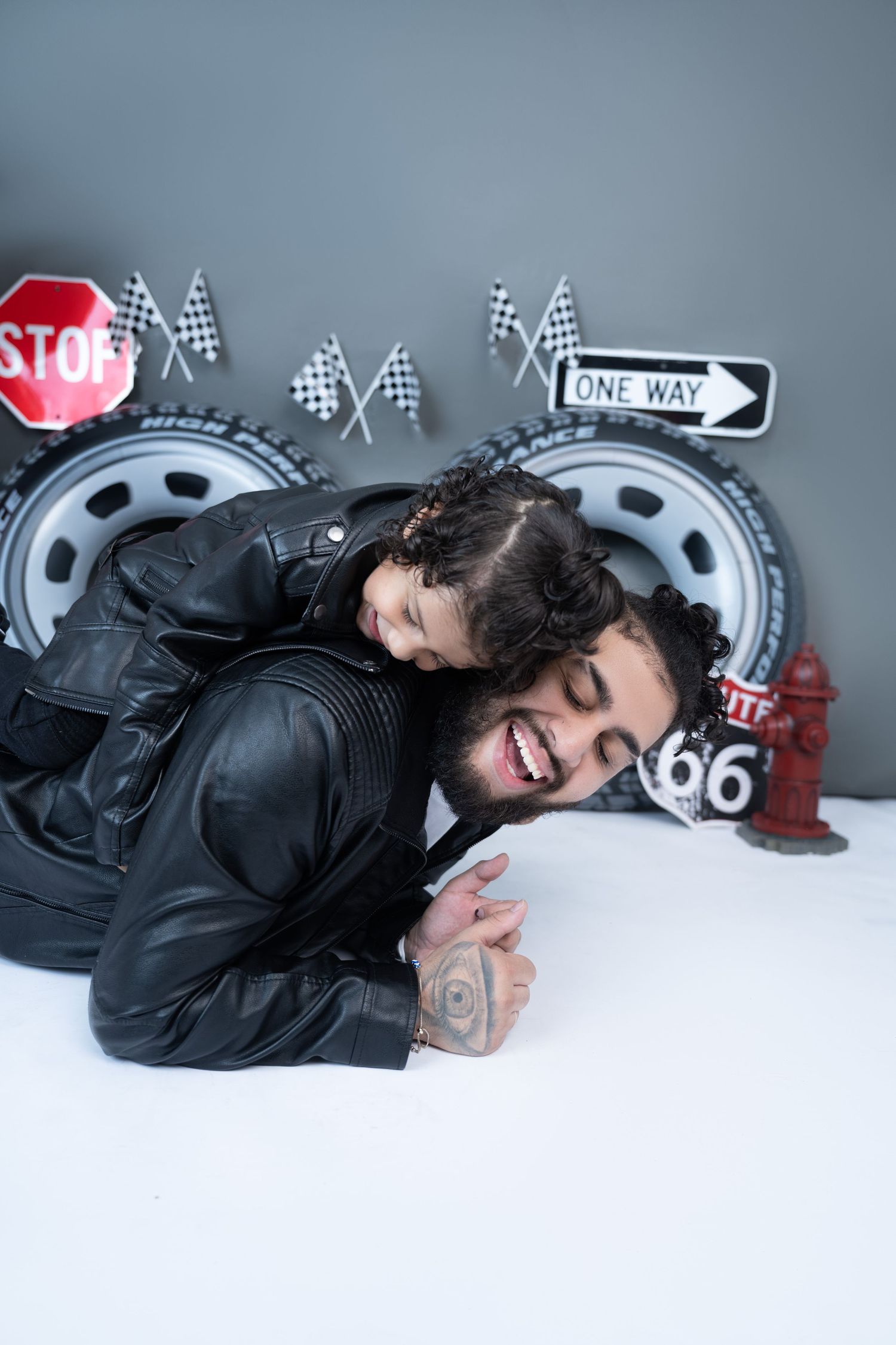 A bearded man in a leather jacket laughs while a child climbs on his back in a racing-themed photo studio.