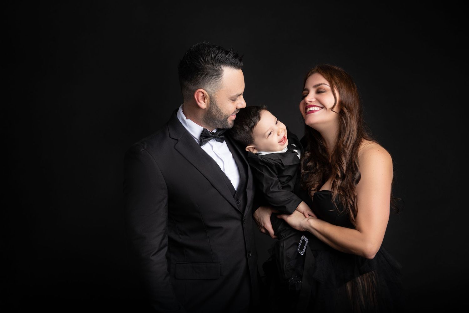 A couple in formal black attire smiling at their baby boy against a dark studio background.