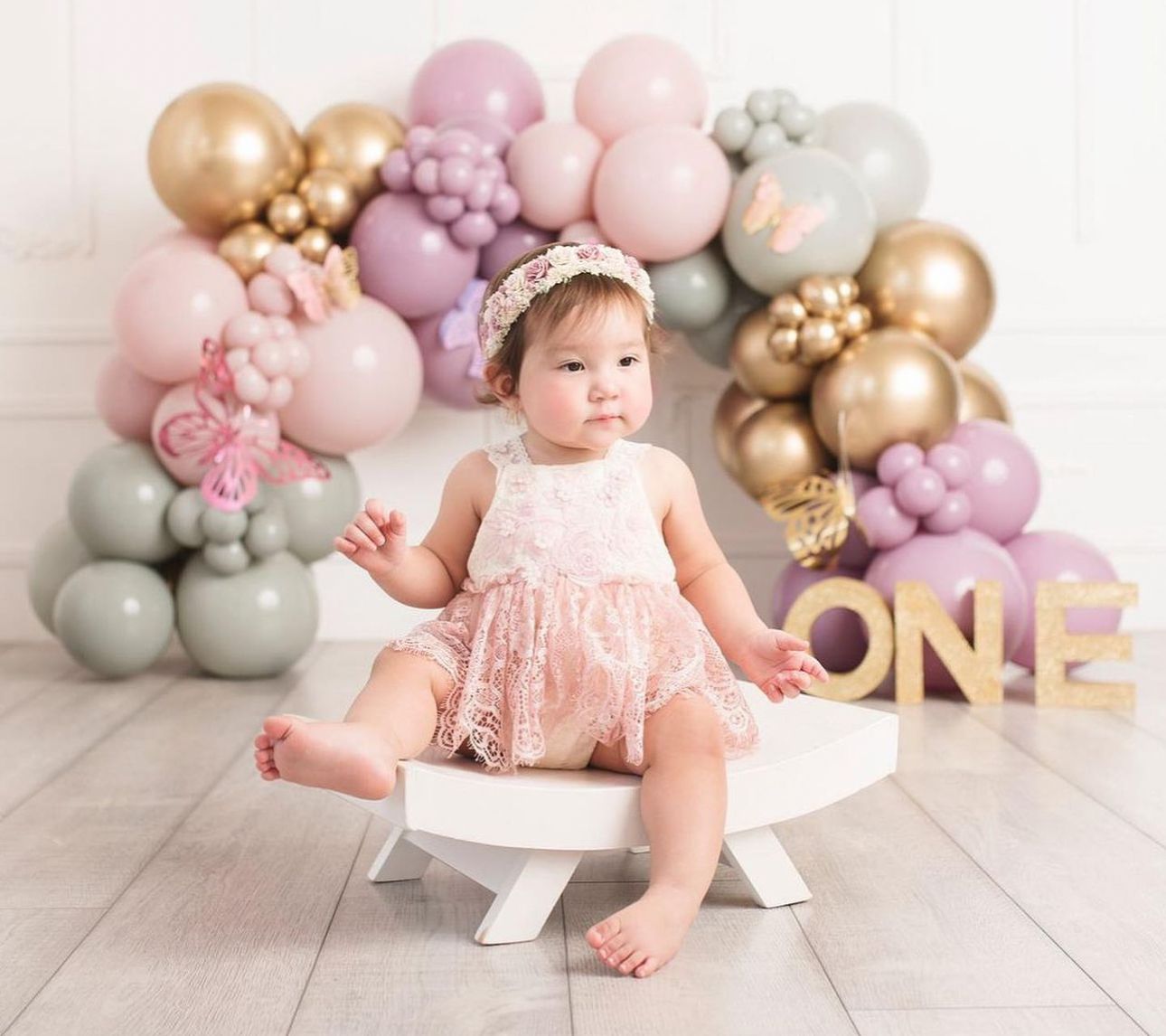 First birthday photo setup with pink dress, gold and pastel balloon arch, and decorative 'ONE' letters on white floor.