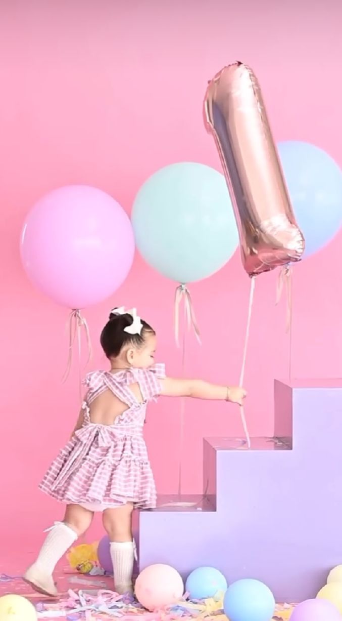 Small child in pink dress climbing purple stairs with pastel balloons against pink backdrop for birthday photoshoot.