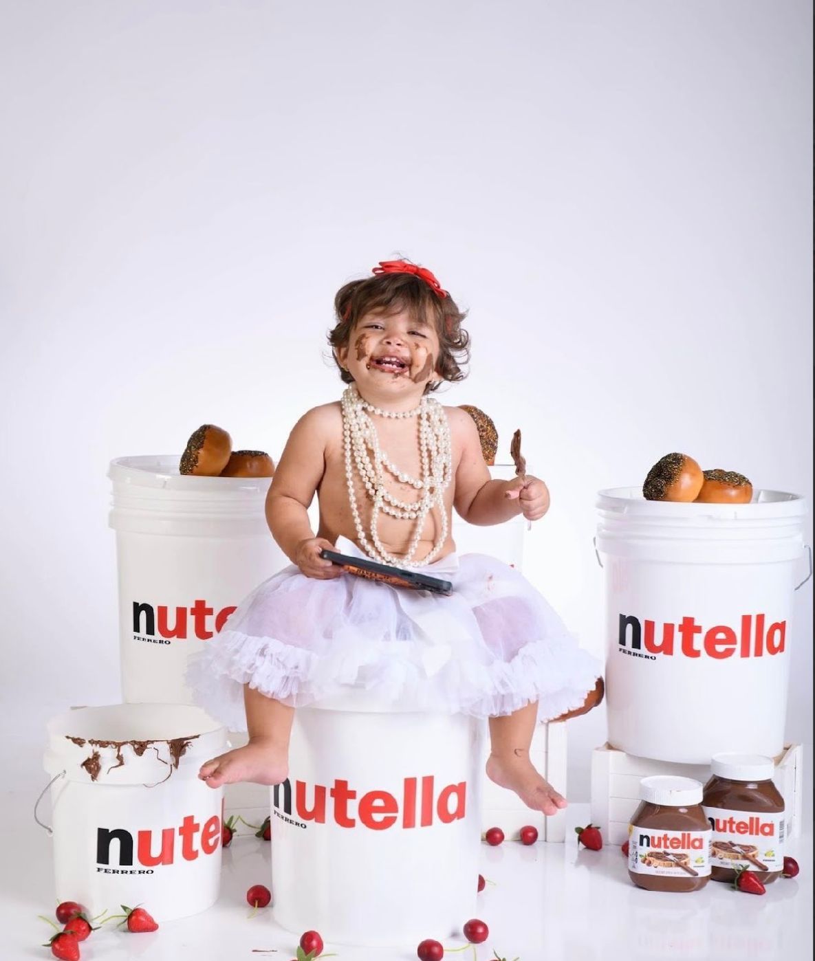 A series of photos showing a baby in a white tutu dress posing with Nutella jars and strawberries on a white backdrop.