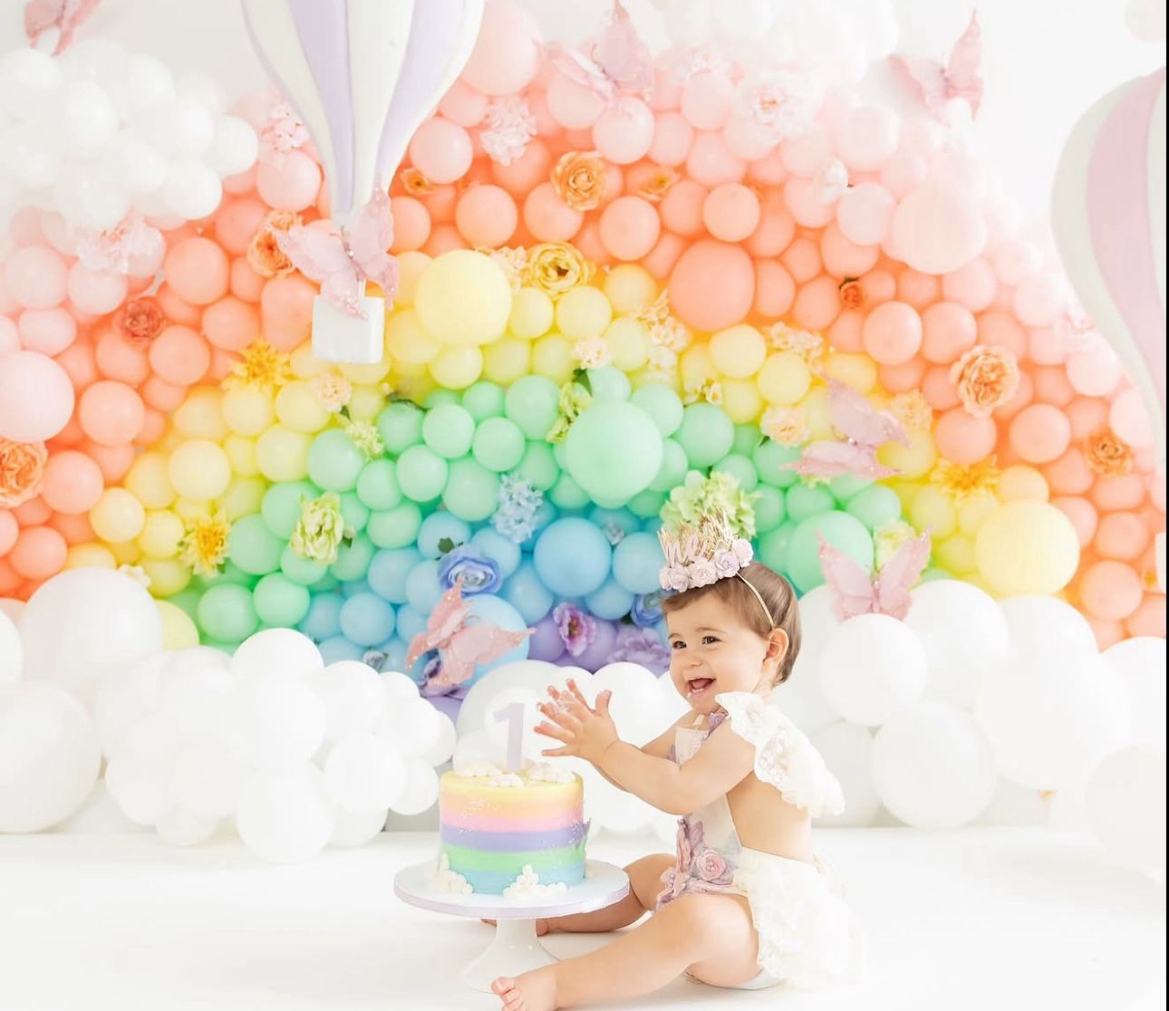 A rainbow balloon arch installation creates a magical backdrop for a baby sitting with an Easter egg in a dreamy setting.