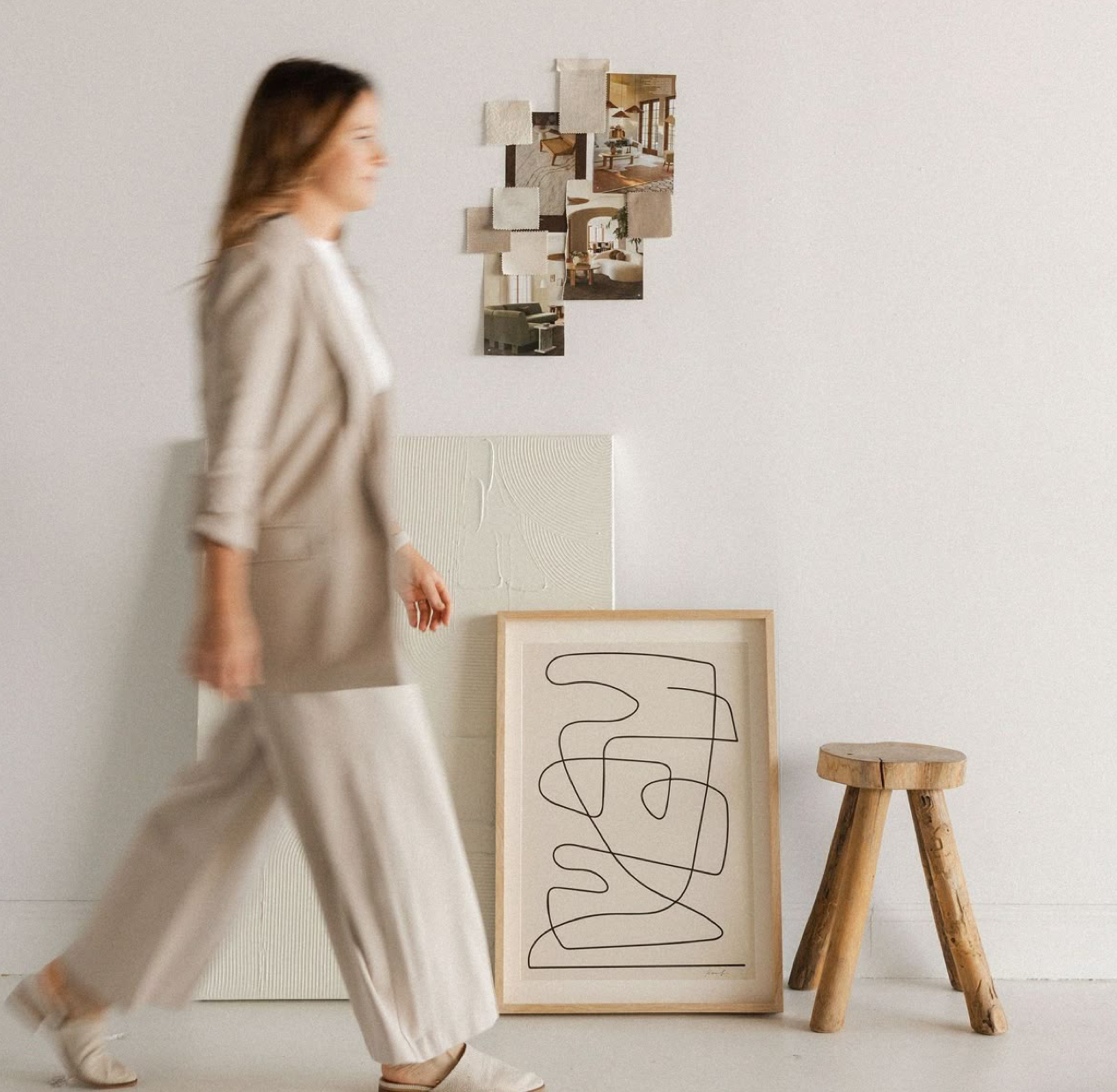 Woman in neutral linen outfit walking past framed abstract line art print and wooden stool in minimalist white room.