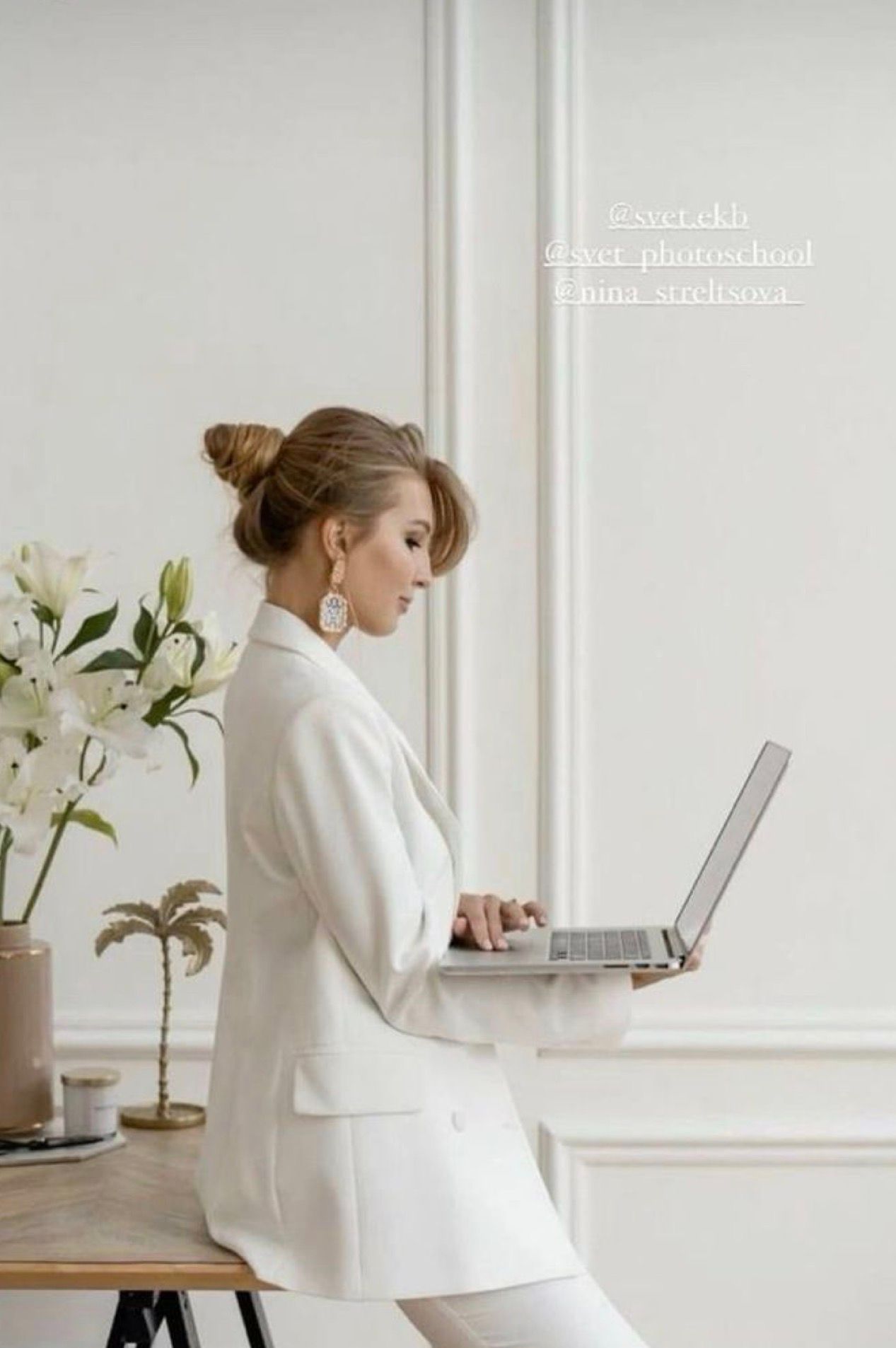 A professional woman in a white blazer suit sits on a desk using a laptop in a bright, elegant white office setting.