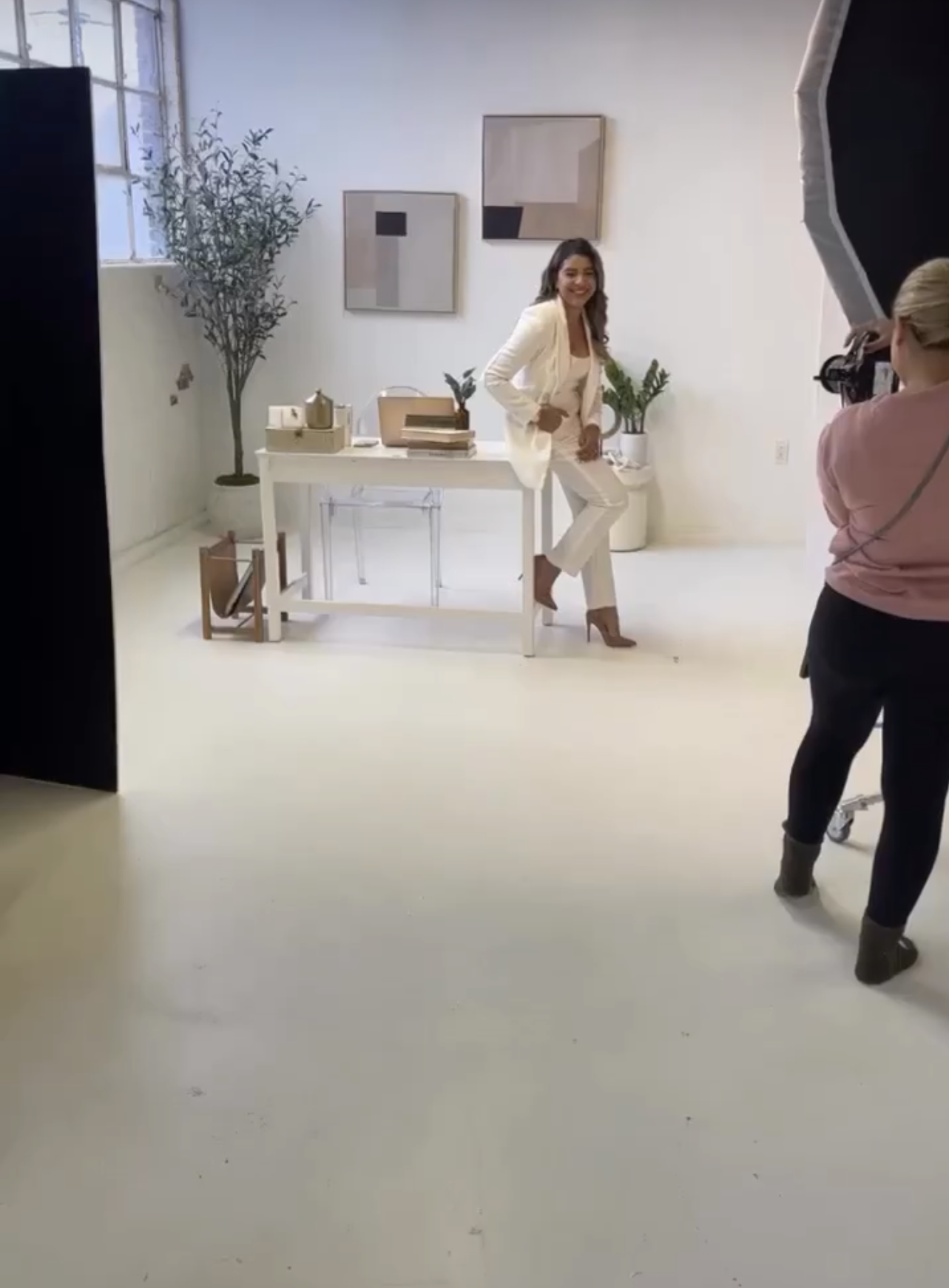 Woman in white suit poses for photographer in bright studio with neutral decor and olive tree backdrop.