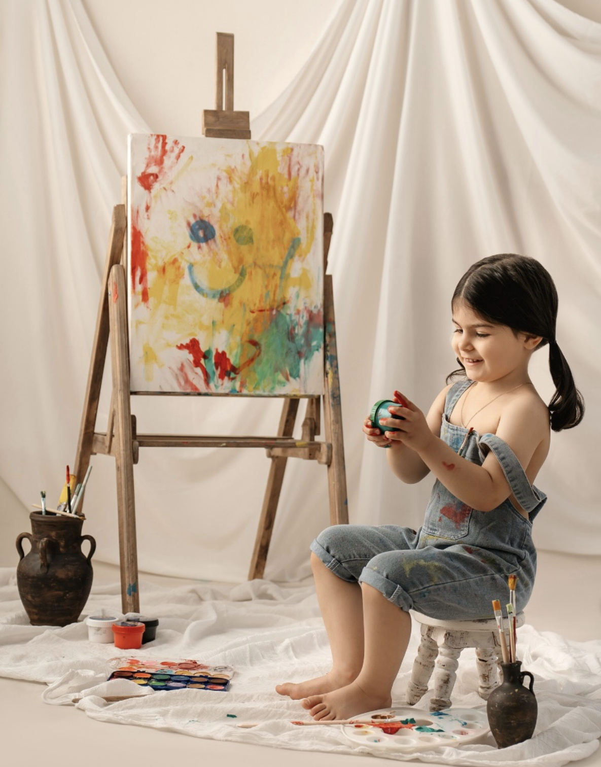 Young girl in denim overalls sits on stool holding painted ball beside colorful canvas on easel with art supplies nearby.