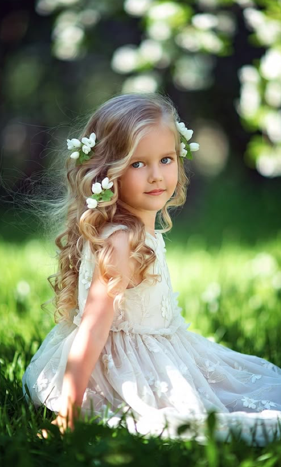 Young girl with curly blonde hair adorned with white flowers wearing a lace dress sitting in green grass outdoors.