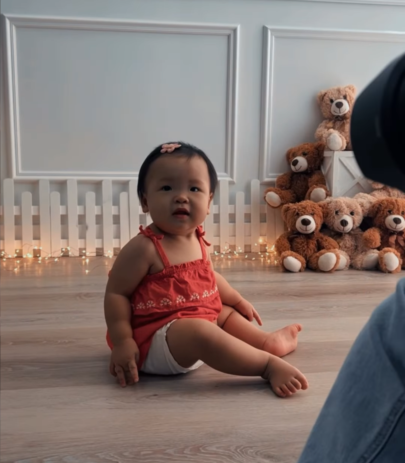 Cute baby girl in red top sits on hardwood floor surrounded by teddy bears and fairy lights.