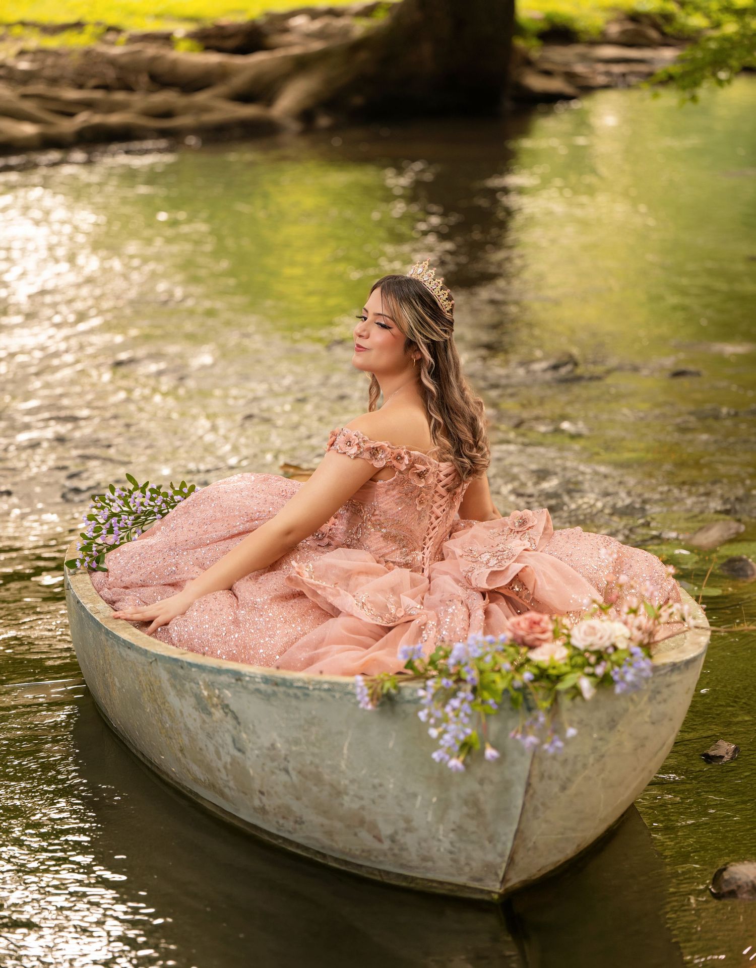 Young woman in pink ball gown sitting in a boat on a serene river, surrounded by flowers.