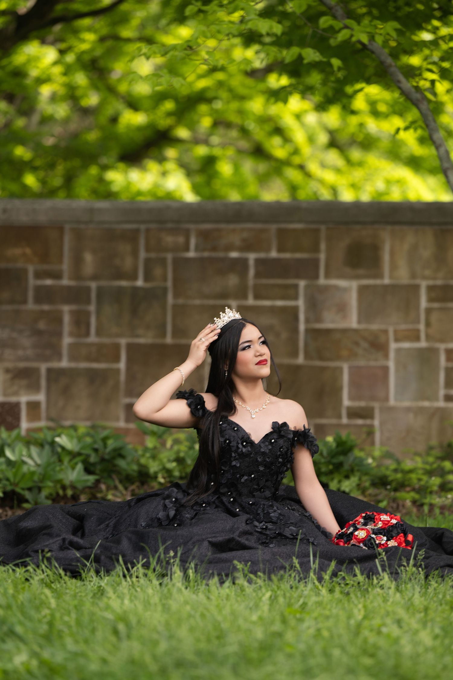 Young woman in black ball gown and tiara sits gracefully on grass before stone wall with lush green trees.