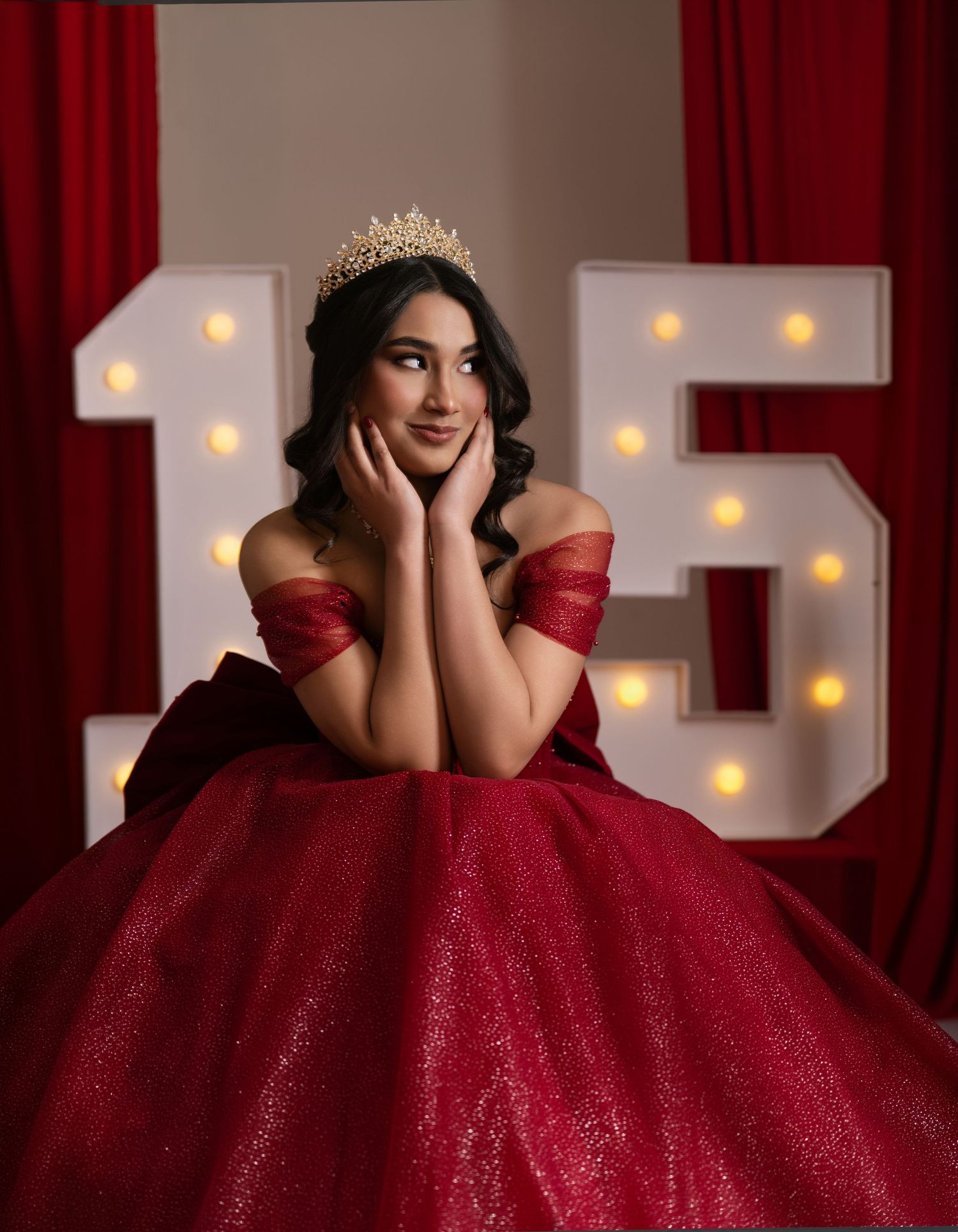 Young woman in red ball gown and gold crown poses before illuminated number 15 marquee letters for quinceañera.