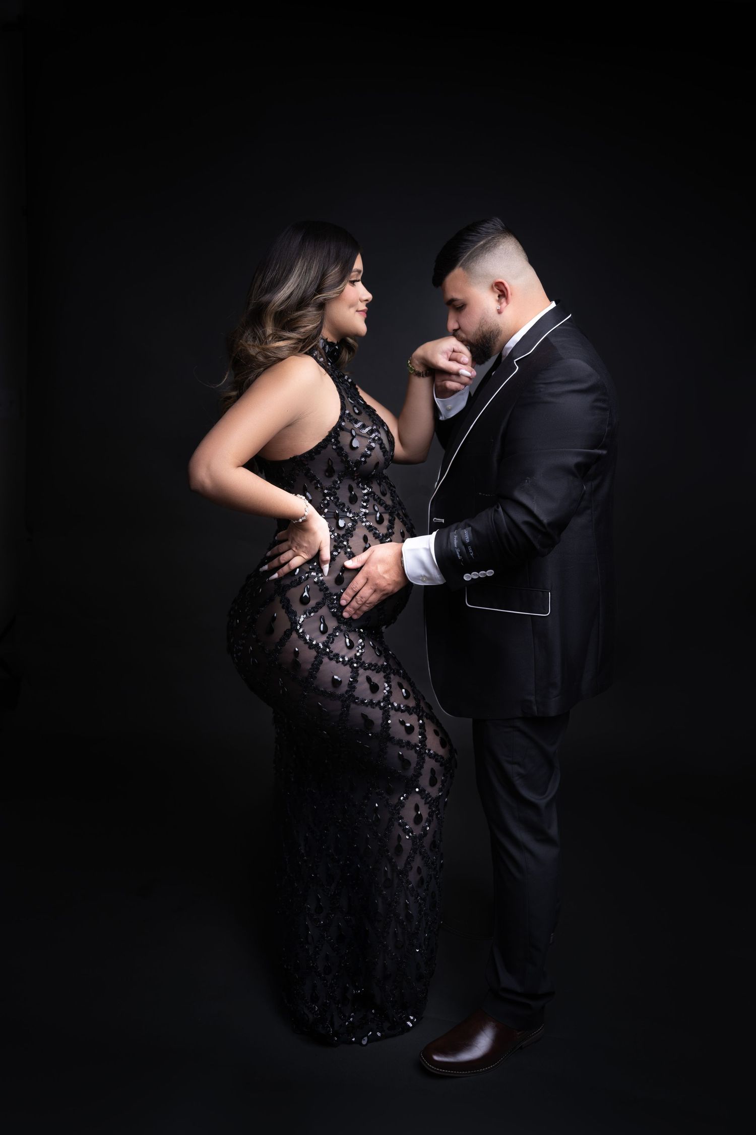 Elegant couple in black formal wear posing together, pregnant woman in lace gown with man in suit kissing her hand.