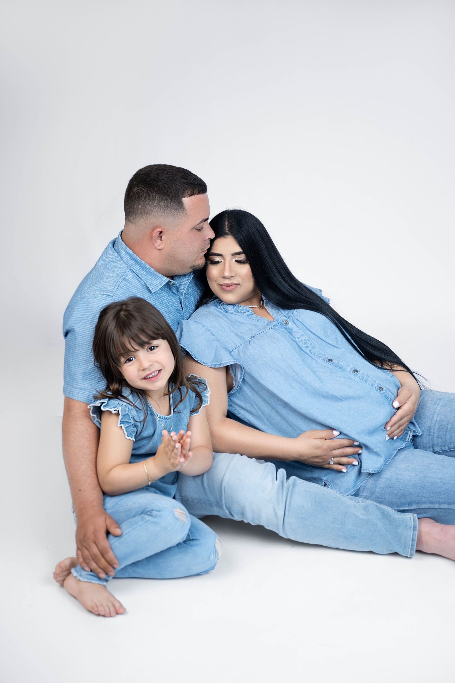 A family in matching denim outfits poses together, with a man kissing a pregnant woman while a young girl smiles.