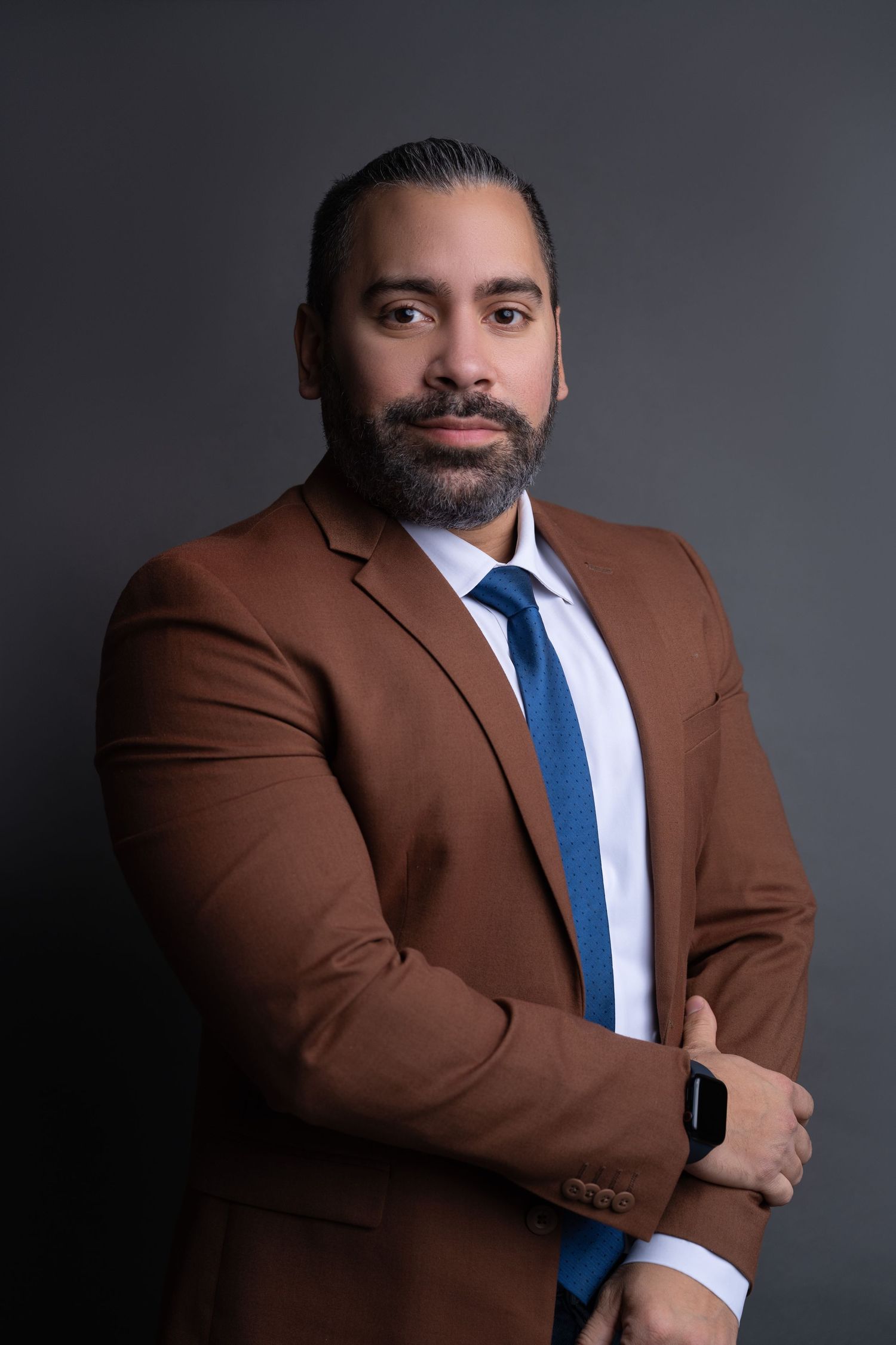 Professional studio portrait of a bearded man in a brown blazer, white shirt and blue tie, arms crossed, wearing a smartwatch.