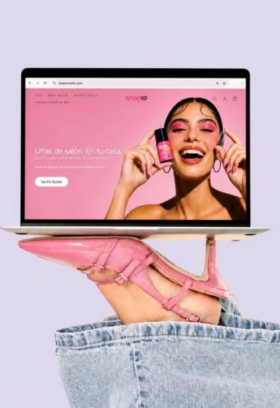 Pink high-heel mule shoe peeking from denim jeans pocket beside a laptop displaying a nail beauty e-commerce website.