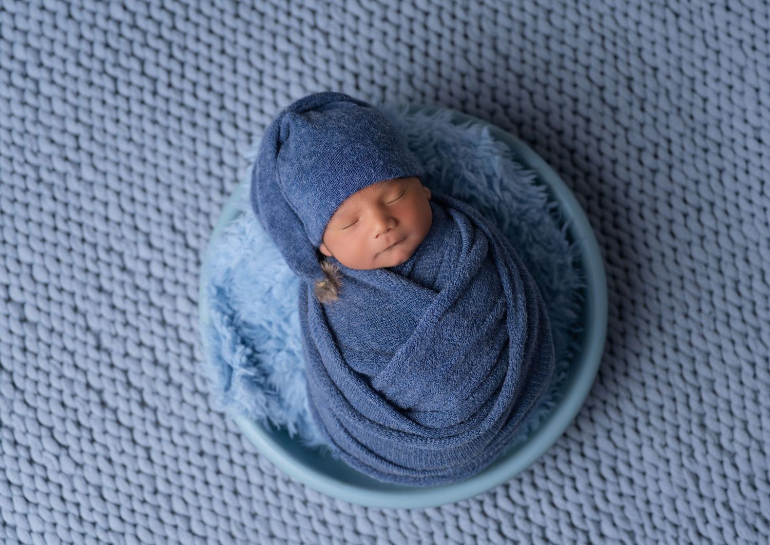 Newborn baby swaddled in blue wrap with matching knit hat, sleeping peacefully in a teal bowl on blue background.